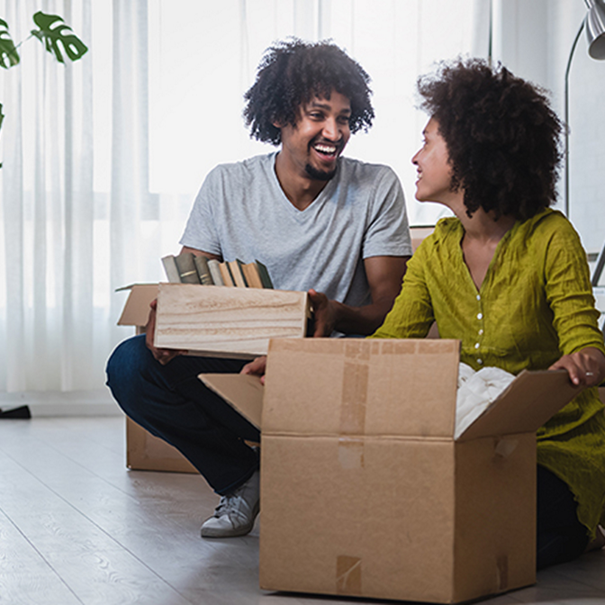 Couple laughing sitting on floor in living room surrounded by moving boxes